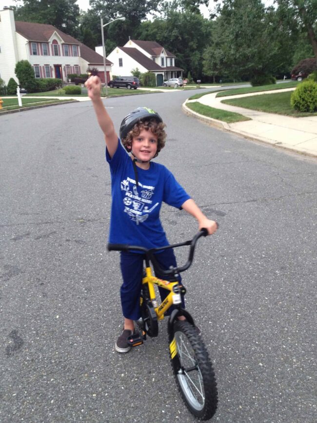 Child riding a yellow bike with training wheels, wearing a helmet, and raising his hand in a suburba.