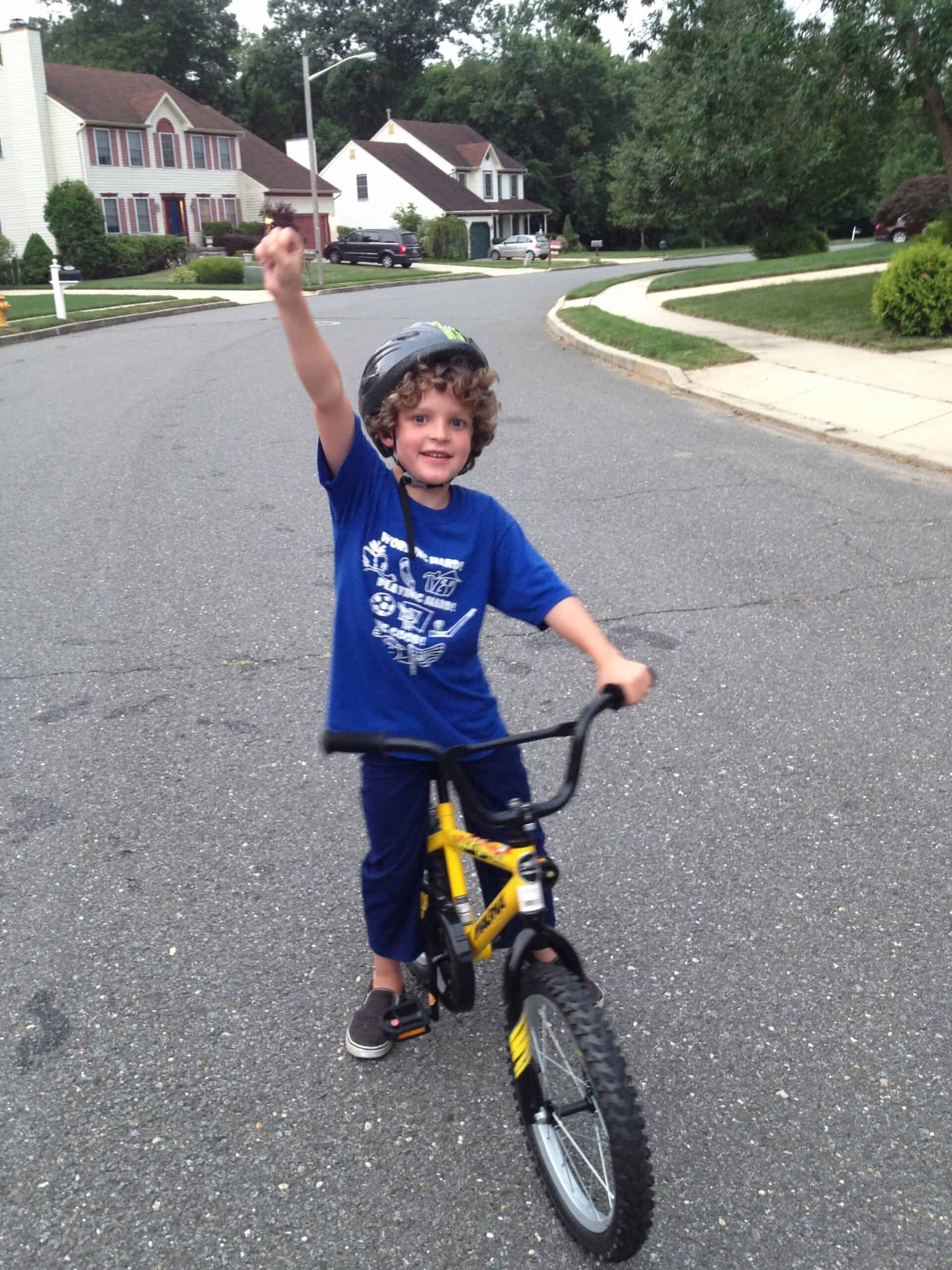 Child riding a yellow bike with training wheels, wearing a helmet, and raising his hand in a suburba.
